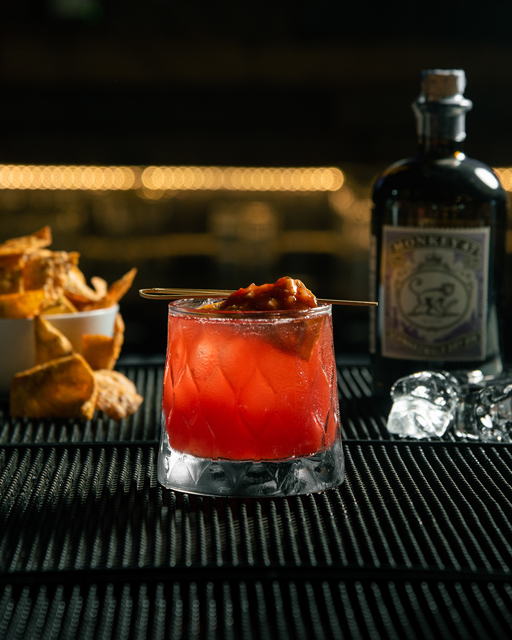 A colorful cocktail garnished with a slice of fruit, accompanied by a small bowl of crispy chips