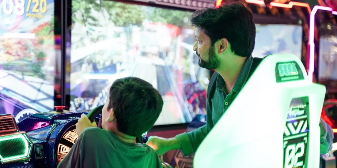 A man and a boy enthusiastically playing video games together in a vibrant arcade filled with colorful machines