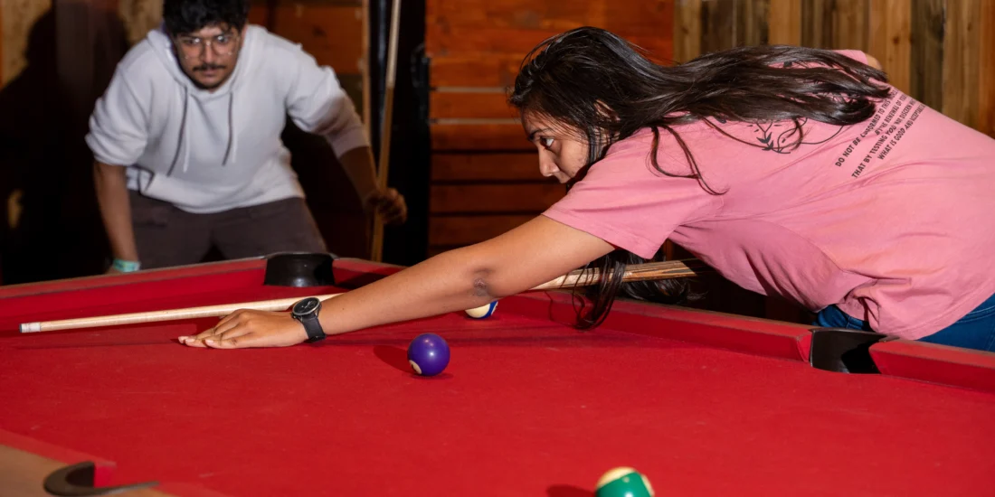 Pink tshirt woment playing 8pool and a men standing near with the white dress
