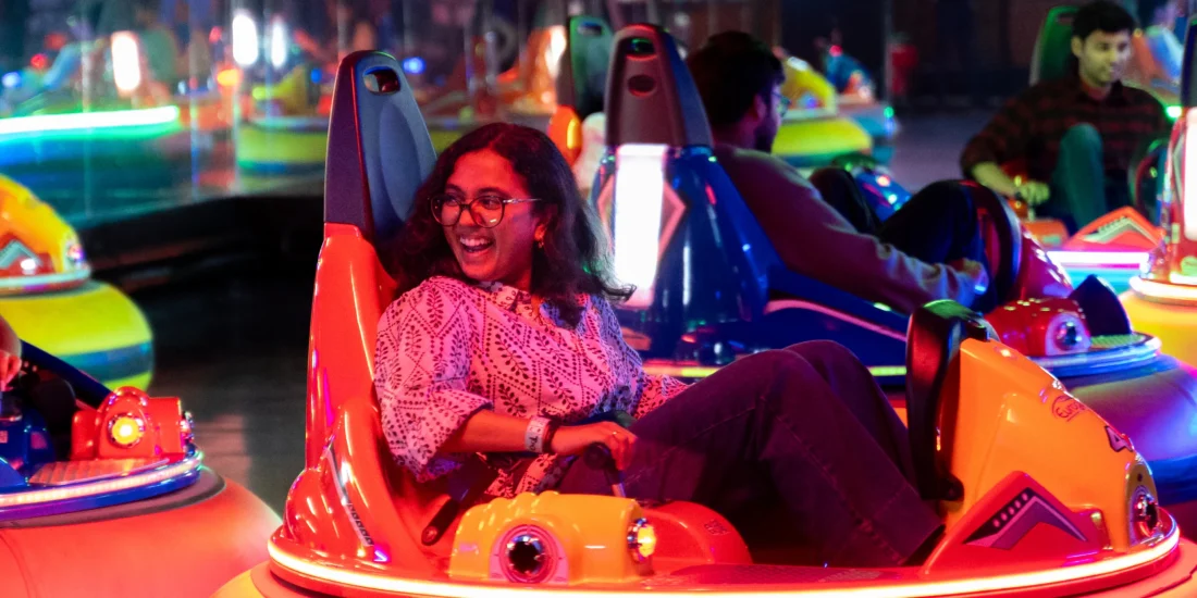 A smiling woman enjoys riding bumper cars at an amusement park, surrounded by colorful lights and joyful atmosphere