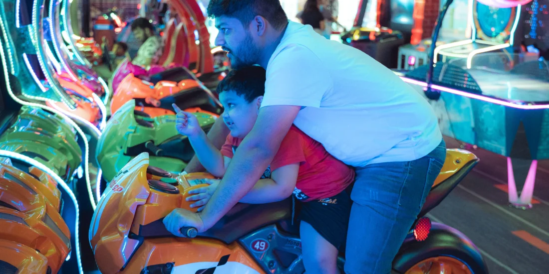 Father and Son enjoy a motorcycle ride in an arcade, surrounded by colorful game machines and bright lights