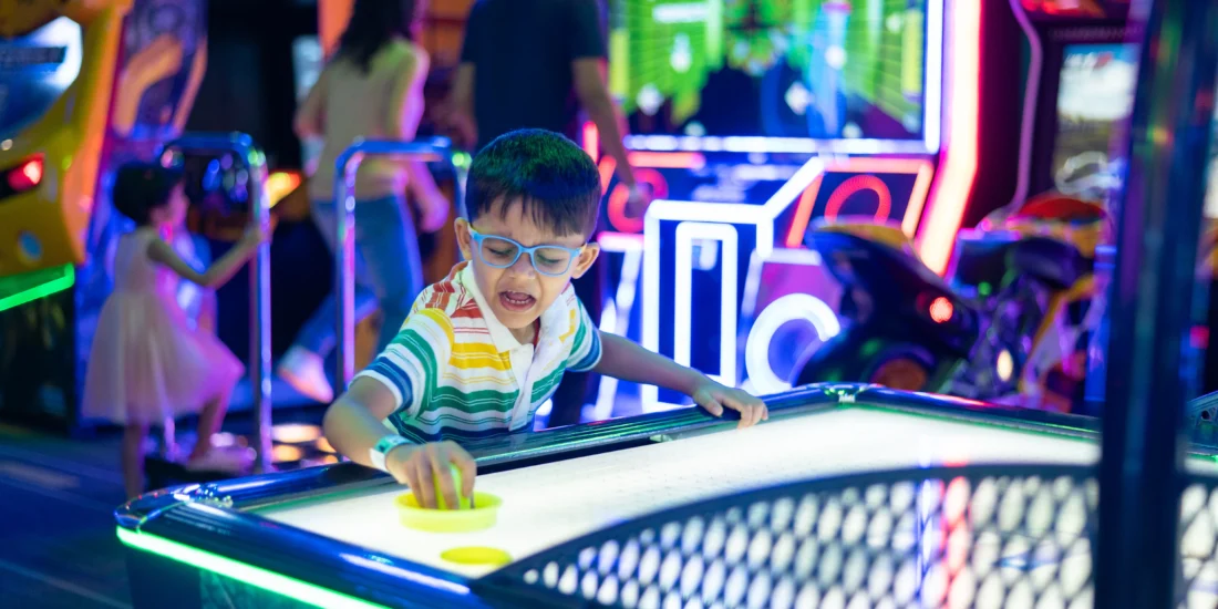 A young boy focused on playing air hockey, striking the puck with enthusiasm at a brightly lit arcade
