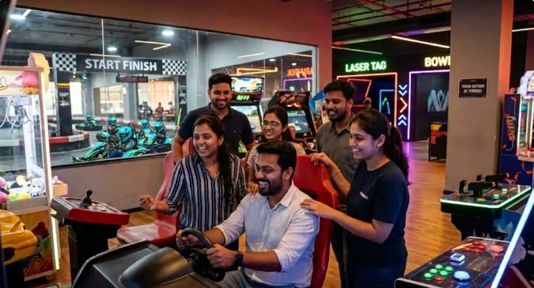 Diverse group of corporate colleagues cheering around a multiplayer racing simulator arcade game at Torq03 Bengaluru, with blue go-karts racing on an indoor track visible through the background glass partition.