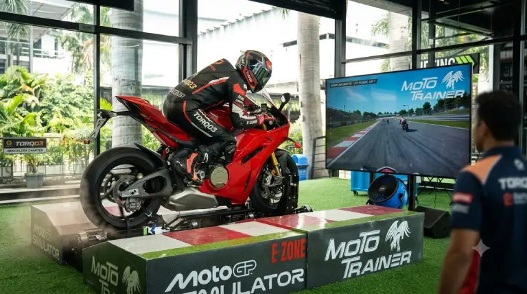A rider fully geared in a leather racing suit and helmet leaning into a corner on a red superbike mounted on the Moto Trainer MotoGP Simulator platform at Torq03 Bangalore, with an instructor observing.